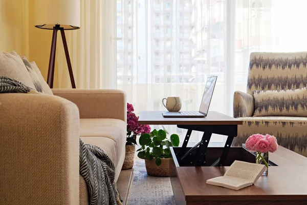 Living room with modern beige textile couch and laptop on wood textured table, natural light. House plants in cozy interior. Background, copy space/