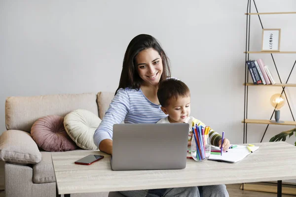 Young beautiful woman multitasking, working from home & teaching two year old son to draw. Woman spending quality time with her toddler child. Homeschooling concept. Close up, copy space, background.