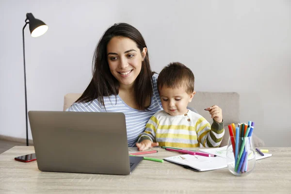 Young beautiful woman multitasking, working from home & teaching two year old son to draw. Woman spending quality time with her toddler child. Homeschooling concept. Close up, copy space, background.