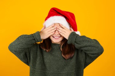 Geleneksel Noel Baba şapkası ve yeşil örgü süveter giyen genç kızın portresi izole edilmiş sarı arka planda poz veriyor. Noel 'i kutlayan bir çocuğun stüdyo fotoğrafı. Kapat, boşluğu kopyala.