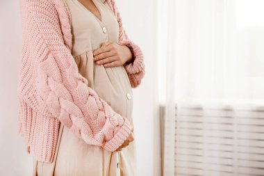 Hamileliğin ikinci üç ayında genç bir kadının kırpılmış görüntüsü. Kolları göbeğinde, pembe büyük örülü süveter giyen hamile kadına yakın durun. Çocuk konsepti bekliyorum. Arkaplan, boşluğu kopyala.