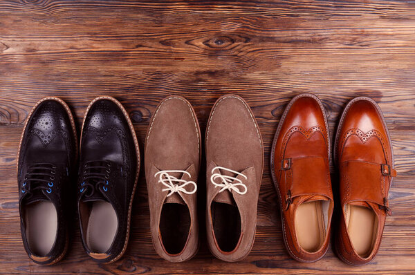 Bunch of different style men's shoes in a row. Close up shot chukka boots, single and double monk strap oxfords, brown, black and burgundy brogues. Top view, copy space, flat lay, wooden background