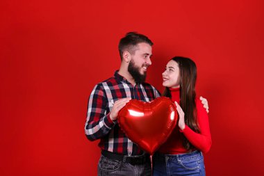 Sevgililer Günü konseptin kutlu olsun. Kalp şeklinde balonla sıradan kıyafetler giyen çiftlerin stüdyo fotoğrafları. Hippi sakallı adam ve kızın portresi sevgi gösteriyor. Kırmızı duvar arkaplanı, alanı kopyala