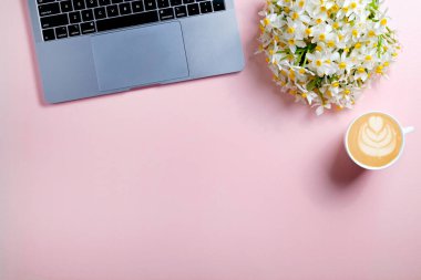 Dizüstü bilgisayarı, bir buket nergis ve bir fincan kapuçino ile kadınsı çalışma alanı. Kadın moda blogcusu konseptinin işyeri. Pembe arkaplan, metin için boşluk kopyala, düz yatay, kapat.