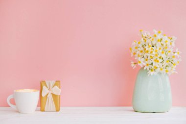 Kaliteli turkuaz vazoda bir buket nergis ve pembe arka planda izole edilmiş bir fincan kapuçino. Yumuşak minimalist bahar çiçekleri kompozisyonu. Üst görünüm, metin için boşluk kopyala, düz görünüm, kapat