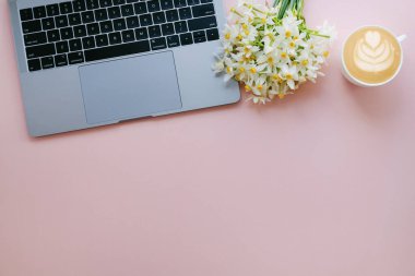 Dizüstü bilgisayarı, bir buket nergis ve bir fincan kapuçino ile kadınsı çalışma alanı. Kadın moda blogcusu konseptinin işyeri. Pembe arkaplan, metin için boşluk kopyala, düz yatay, kapat.