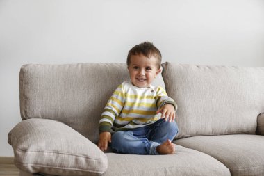 Tekstil kanepesinde oturan ve gülümseyen sevimli küçük çocuğun portresi. Mutlu çocuk evde gülüp eğleniyor. Kot pantolonlu çıplak ayaklı çocuk. Kapat, kopyala boşluk, arkaplan.