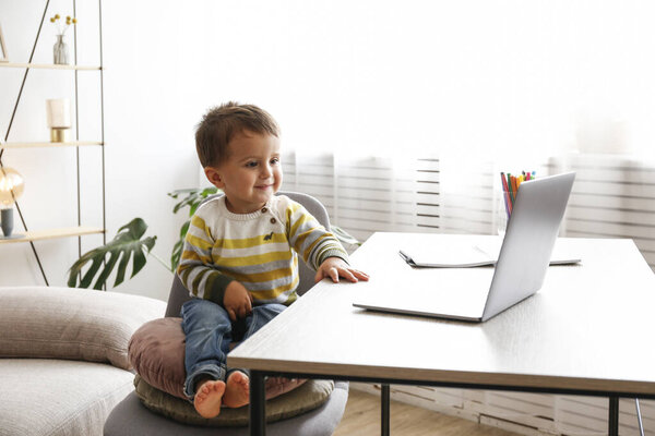 Двухлетний мальчик смотрит на экран ноутбука с очаровательным выражением лица. Маленький мальчик сидит за столом дома с ноутбуком. Закрыть, скопировать пространство, фон.