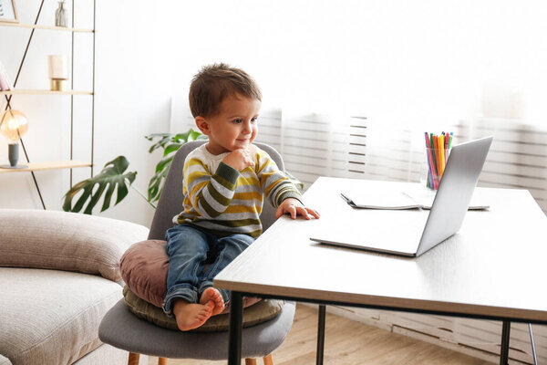 Двухлетний мальчик смотрит на экран ноутбука с очаровательным выражением лица. Маленький мальчик сидит за столом дома с ноутбуком. Закрыть, скопировать пространство, фон.