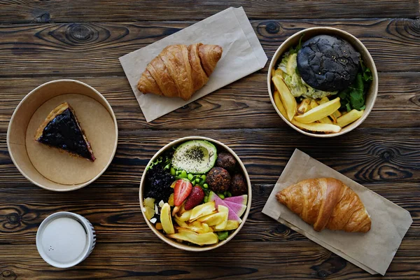 Different takeout food on wooden kitchen table. Hawaiian vegan poke bowl with variety of veggies, french croissant, salmon burger and blueberry cheesecake. Close up, top view, copy space, background.