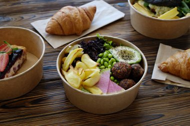 Ahşap mutfak masasında farklı servis yemekleri. Çeşitli sebzeler, Fransız kruvasanı, somon burger ve yabanmersinli cheesecake ile Hawaii vejetaryen poke kasesi. Kapat, üst görünüm, kopyalama alanı, arkaplan.