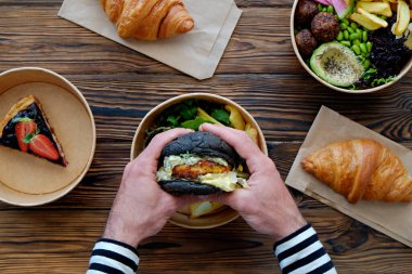 Ahşap masanın üzerinde somon burger ve siyah çörek tutan bir adam resmi. Paket yemek. Fransız kruvasanı, Hawaii vejetaryen dürtme kasesi, yabanmersinli cheesecake. Kapat, üst görünüm, kopyalama alanı, arkaplan