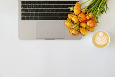 Kırpılmış çekim ya da dizüstü bilgisayar klavyesi, köpük resimli bir fincan kapuçino ve dokuma masasında yazı için bir sürü yeri olan sarı-turuncu Ranunculus çiçekleri. Kapat, üst görünüm.