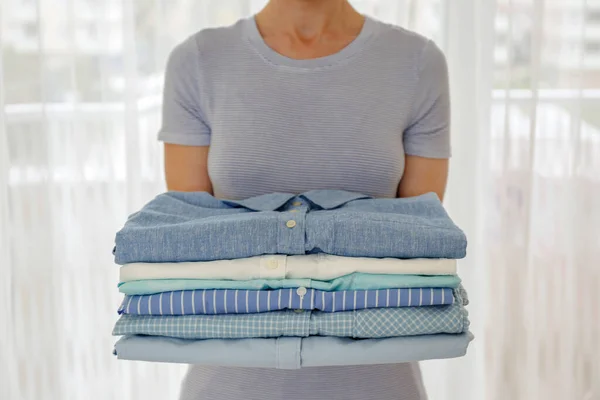 Cropped shot of young woman at home holding stack of perfectly folded shirts. Unrecognizable female with pile of different clothing in her hands. Laundry day concept. Close up, copy space, background