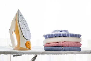 Evde temiz, yeni yıkanmış giysiler ve elektrikli ütü yığını var. Ütü masasında bir yığın gömlek, beyaz perdeli uzun bir pencere. Boşluğu kopyala, kapat, üst görünüm, arkaplan