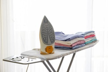 Evde temiz, yeni yıkanmış giysiler ve elektrikli ütü yığını var. Ütü masasında bir yığın gömlek, beyaz perdeli uzun bir pencere. Boşluğu kopyala, kapat, üst görünüm, arkaplan