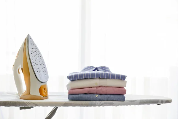 Stack of clean freshly laundered clothes and electric iron on pressboard at home. Pile of shirts on ironing board, full length window with white curtains. Copy space, close up, top view, background