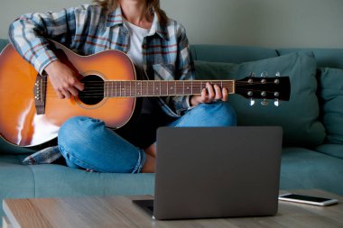 Kareli tişörtlü müzik koleji öğrencisi akustik gitar egzersizi yapıyor, dizüstü bilgisayardan notlar okuyor. Genç bir kadın çevrimiçi müzik dersleri alıyor, evde pratik yapıyor. Arkaplan, kapat