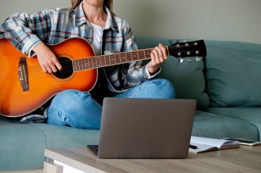 Kareli tişörtlü müzik koleji öğrencisi akustik gitar egzersizi yapıyor, dizüstü bilgisayardan notlar okuyor. Genç bir kadın çevrimiçi müzik dersleri alıyor, evde pratik yapıyor. Arkaplan, kapat