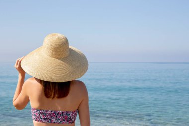 Çiçek desenli parlak bikinili ve geniş kenarlı hasır şapkalı çekici genç bir kadının sahilde dikilip okyanus manzarasının keyfini çıkardığı arka plan görüntüsü. Arkaplan, kapat, boşluğu kopyala