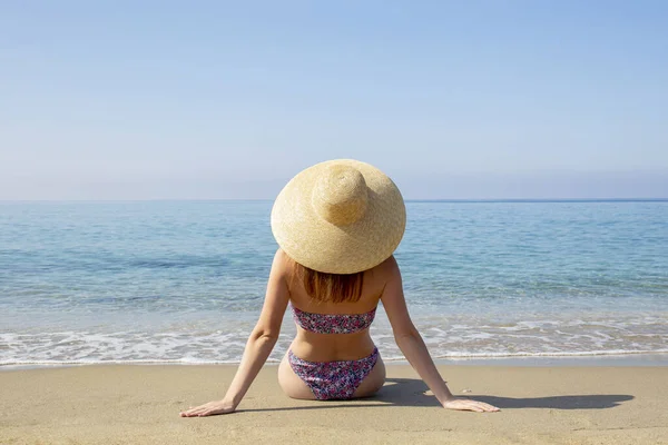 魅力的なフィット感のバックビュー花のパターンと広いつばのわらの帽子と明るいビキニ水着を身に着けている若い女性は 海の景色を楽しんで 砂浜に座っている コピースペース — ストック写真