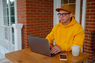 Parlak hardal sarısı kapüşonlu genç adam kahve dükkanının avlusunda dizüstü bilgisayarıyla çalışıyor. Elektronik aletleri ve kişisel kupası olan hippi adam. Kapat, kopyala boşluk, arkaplan.