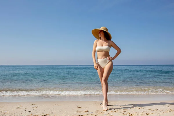 Caucasian woman with fit body bikini bathing suit with high waisted bottom and broad brim straw hat posing at sandy beach on beautiful sunny day. Mediterranean sea background. Copy space