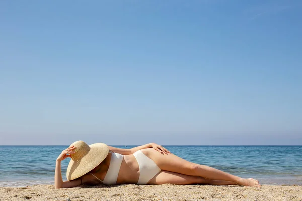 高い腰底と広いつばの藁帽子を持つフィットボディビキニ水着を持つ白人女性は美しい晴れた日に砂浜でポーズをとっています 地中海の海の背景 スペースのコピー — ストック写真