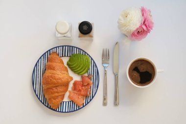 Füme somonlu Fransız kruvasanı, çizgili seramik tabakta avokado, bir fincan sade kahve ve ranunculus çiçeği. Kapat, üst görünüm, düz görünüm, izole edilmiş, metin için boşluğu kopyala.