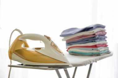 Temiz, yeni yıkanmış giysiler ve elektrikli ütü evdeki basın panosunda. Ütü masasında bir yığın gömlek, tam boy pencere arkası. Boşluğu kopyala, kapat, üst görünüm.