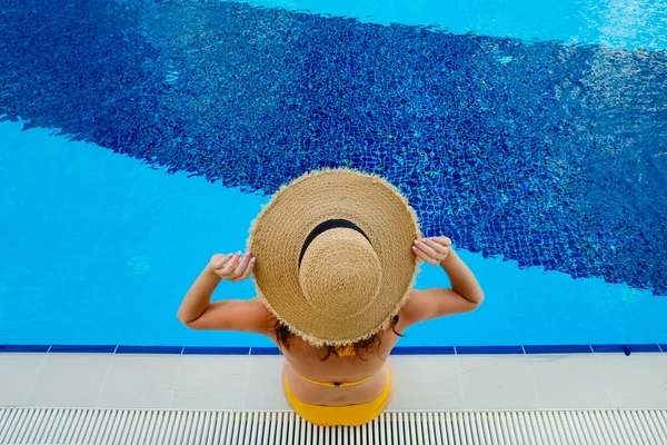Unrecognizable caucasian woman with fit body wearing yellow bikini bathing suit and broad brim straw hat covering her face, chilling in a swimming pool. Copy space, top view.