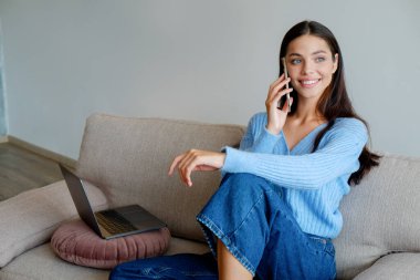 Neşeli kadın eğlenceli bir telefon görüşmesi yapıyor. Kot pantolon giyen esmer bir kadının portresi. Evdeki kanepede oturup gülümsüyor. Arkaplan, kopyala boşluk, kapat.
