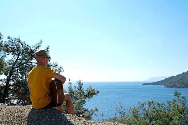 ギターを持って急な丘の上に座って明るい黄色のTシャツを着ている若い男 地中海の島々の美しい景色を楽しむ男性観光客 コピースペース — ストック写真