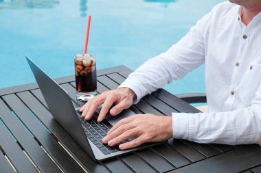 Beyaz keten gömlekli genç adam havuz başında dizüstü bilgisayarıyla çalışıyor. Klavyede yazan erkek seyahat blogcusu. Serbest çalışmanın faydaları işte. Kapat, kopyala boşluk, arkaplan.