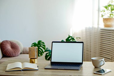 Boş ekran laptop, cep telefonu ve ahşap masada bir fincan. Doğal güneş ışığıyla dolu dekoratif palmiye bitkileriyle dolu rahat bir ofis. Kapat, kopyala boşluk, arkaplan.