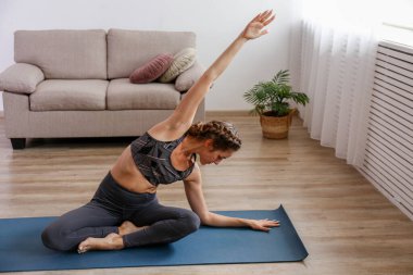 Spor yapan genç bir kadın kendi evinde yoga yapıyor. Yogini oturma odasında sabah egzersizi yapıyor. İç arkaplan, kopyalama alanı, kapat.
