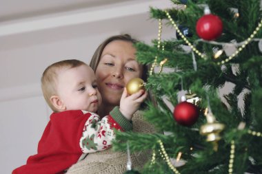 Annem ve yeni doğan bebeği birlikte Noel ağacı süslüyorlar. Boşluğu kopyala, arkaplan.