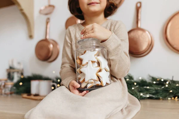 Bej örülü elbise giyen küçük bir kız elinde Noel kurabiyesi şeklinde cam bir kavanoz tutuyor. Minimalist tatil sezonu konsepti