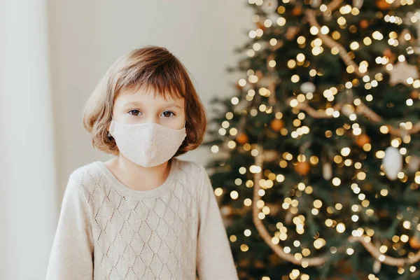 Sad girl meets Christmas in the covid-19 pandemic. Child in a protective medical mask. Quarantine isolation social distance. New year holidays 2021. Kid in Face cover