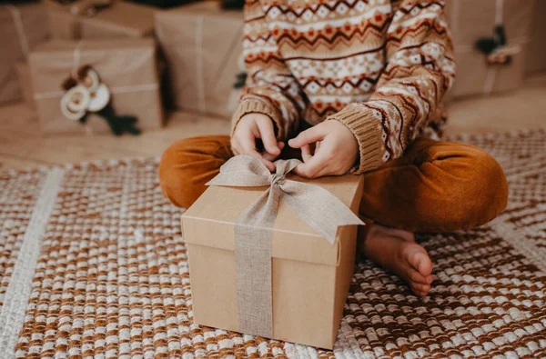 Sıcak süveter giyen çocuk Noel ağacının altında oturuyor ve elinde Noel hediyesi tutuyor. Hediye kutusuna odaklanmış resmi kapat