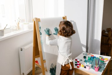 Önlük giyen kız çocuğu çocuk odasındaki sehpada parmak izi bırakıyor. Çocuklar için parmak boyama ya da sanat terapisi. Yürümeye başlayan çocuklar için eğlenceli aktiviteler.