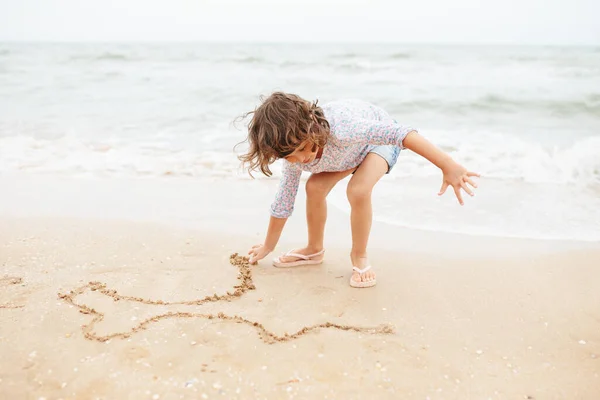 Cinco años lindo rizado caucásico chica pintura en arena en la playa. —  Fotos de Stock