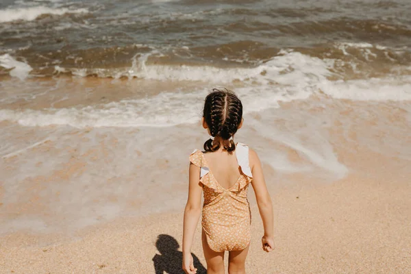 Niña de seis años en traje de baño amarillo de pie en la playa, la gente por detrás. —  Fotos de Stock