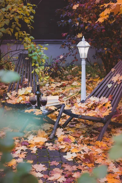 Fall themed calm garden holiday table arrangement with two glasses of red, books, maple leaves, lantern, wooden armchairs, toned