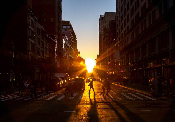 Crowds of people walk through the busy intersection at 23rd Street and Fifth Avenue in New York City with the bright light of sunset casting long shadows on the ground