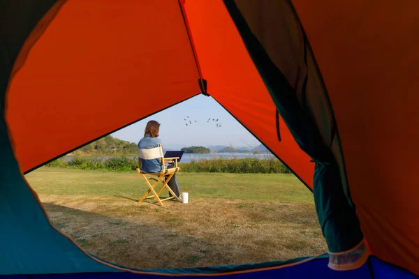 A woman sitting outside a camping tent and using laptop notebook computer working beside wide and big lake with clear blue sky and bids flying in natural scene with coffee cup. Taken from tent inside.