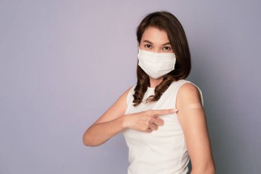 Aşılanmış Asyalı kadın Covid-19 aşısı yaptırdıktan sonra kolunda alçı bandaj gösteriyor ve tıbbi hijyen maskesi takıp otururken gülümsüyor. Coronavirus aşı kampanyası konsepti.