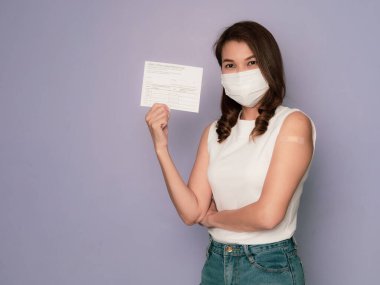 Kollarında yara bandıyla tıbbi koruyucu maske takan ve Coronavirus aşılama kampanyası aşısı olduktan sonra aşılı kayıt kartı gösteren bir kadın..