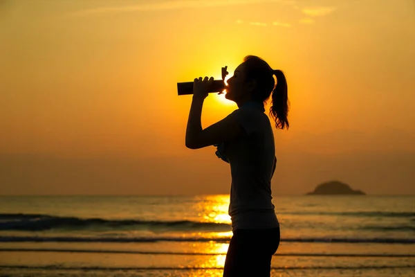 女人站在海滩上 从瓶子里喝着水 做了一个又一个的运动 背景是五彩缤纷的日出 — 图库照片