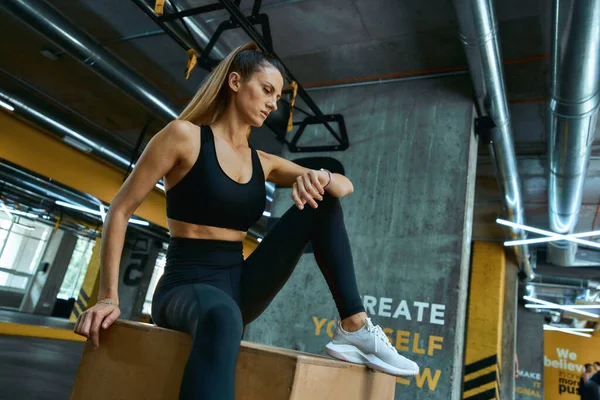 Checking fitness results. Young beautiful athletic girl in sportswear sitting on wooden crossfit jump box at gym and looking at her smartwatch